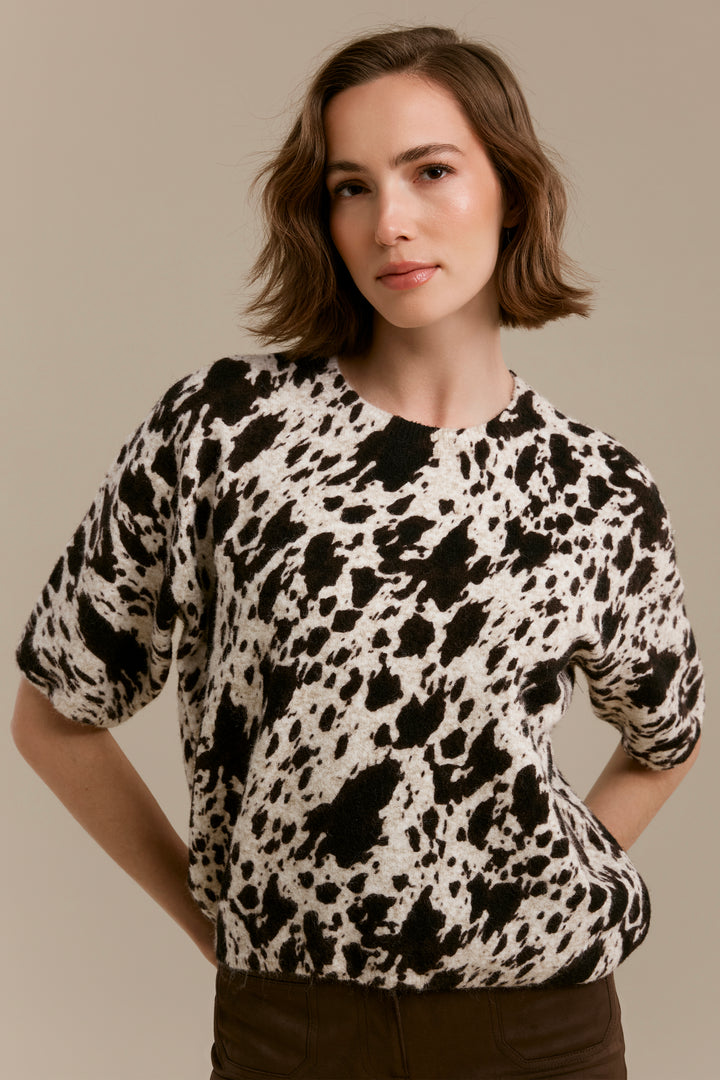 A woman wearing a short-sleeve knit top with a black and off-white cow-print pattern, paired with dark brown pants, against a solid beige background.