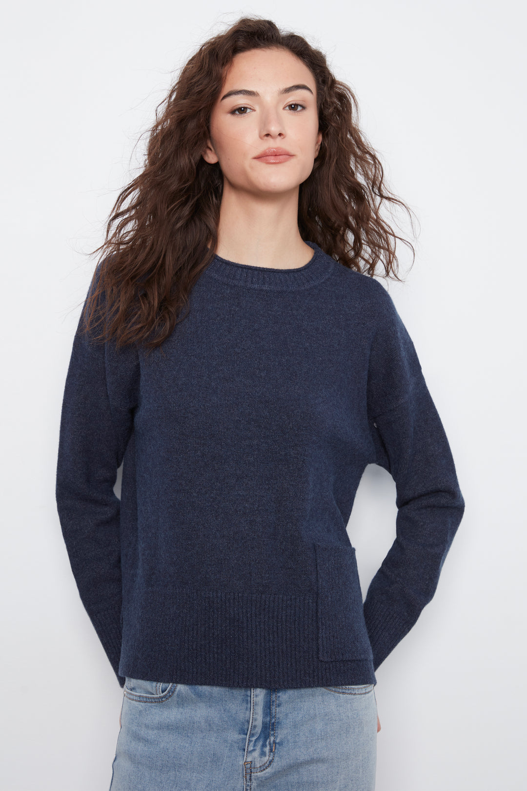 A woman with long, wavy brown hair wearing a dark navy blue crewneck sweater and light wash blue jeans, standing against a plain white studio background.