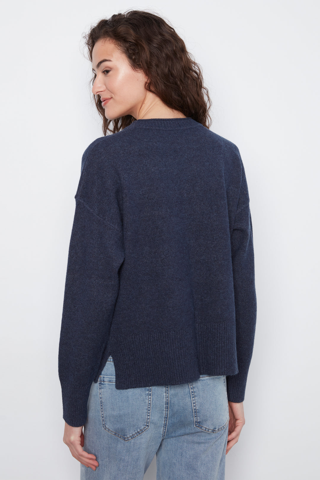 A woman shown from the back wearing a dark navy blue knit sweater and light blue jeans against a plain white studio background.