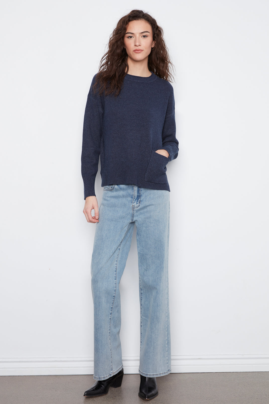 A woman modeling a dark navy blue knit sweater with light-wash wide-leg jeans and black ankle boots, standing against a plain white wall.