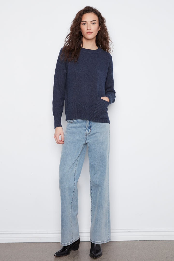 A woman modeling a dark navy blue knit sweater with light-wash wide-leg jeans and black ankle boots, standing against a plain white wall.