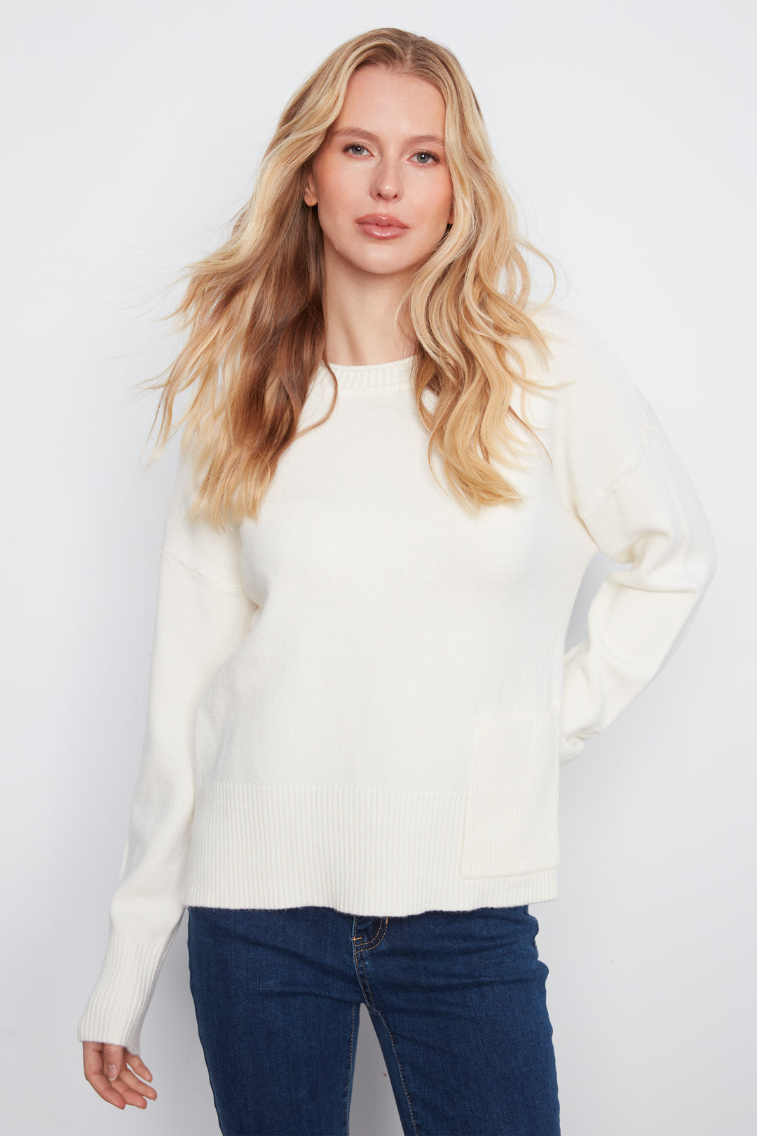 A woman wearing a long-sleeve crewneck sweater in cream with a front patch pocket, paired with blue jeans, against a light gray studio background.
