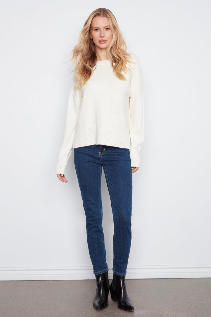 A woman wearing a long-sleeve crewneck sweater in cream with a front patch pocket, paired with blue jeans, against a light gray studio background.