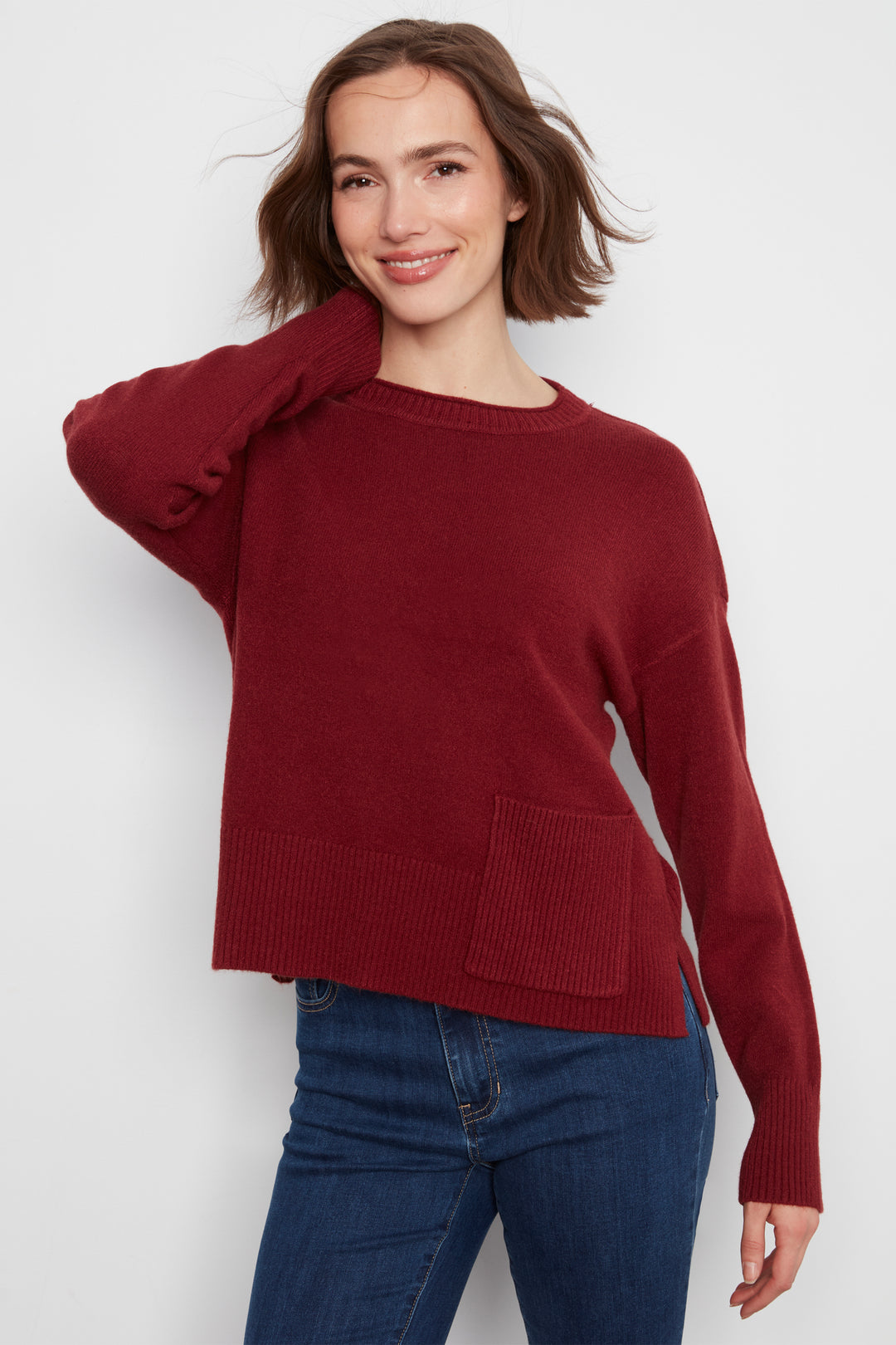 A woman wearing a long-sleeve crewneck sweater in deep burgundy with a front patch pocket, paired with blue jeans, against a light gray studio background.