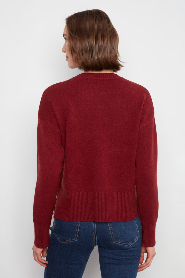 A woman viewed from the back wearing a burgundy knit sweater and blue jeans against a plain light gray studio background.