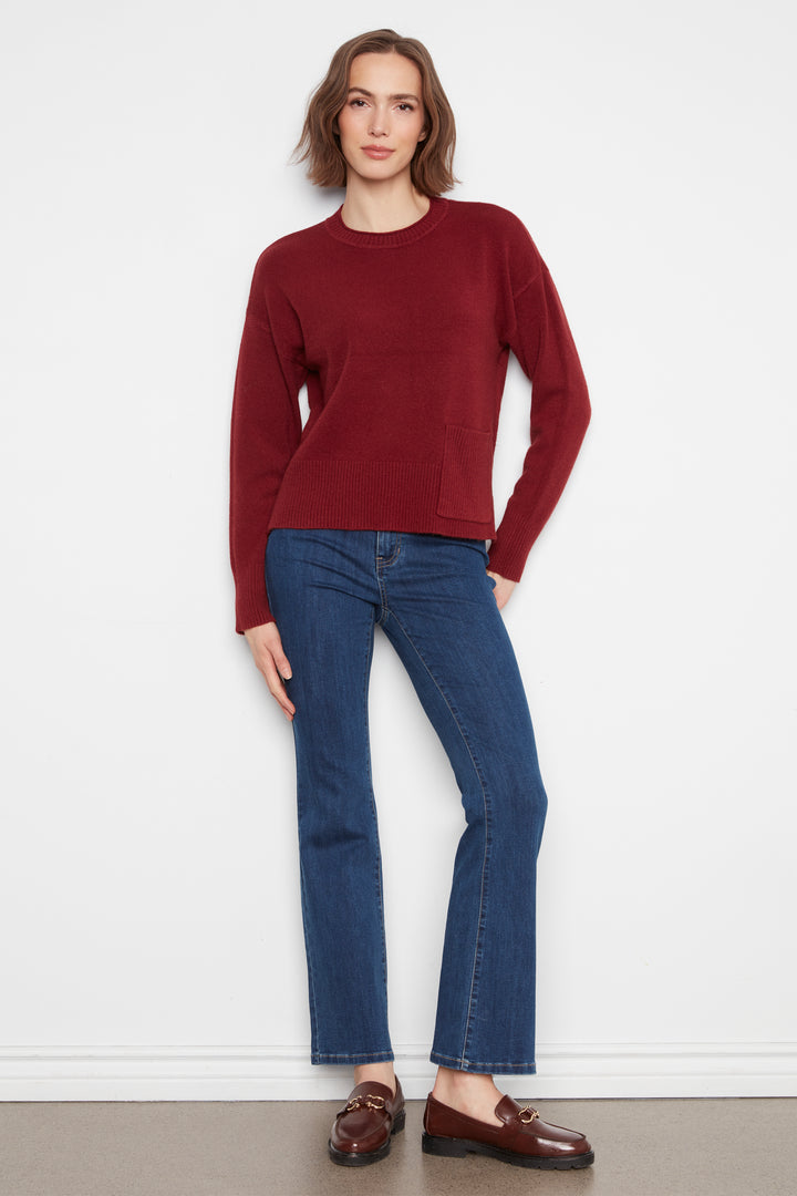 A woman stands against a plain white wall wearing a long-sleeve crewneck sweater in deep burgundy, blue jeans, and brown leather loafers.
