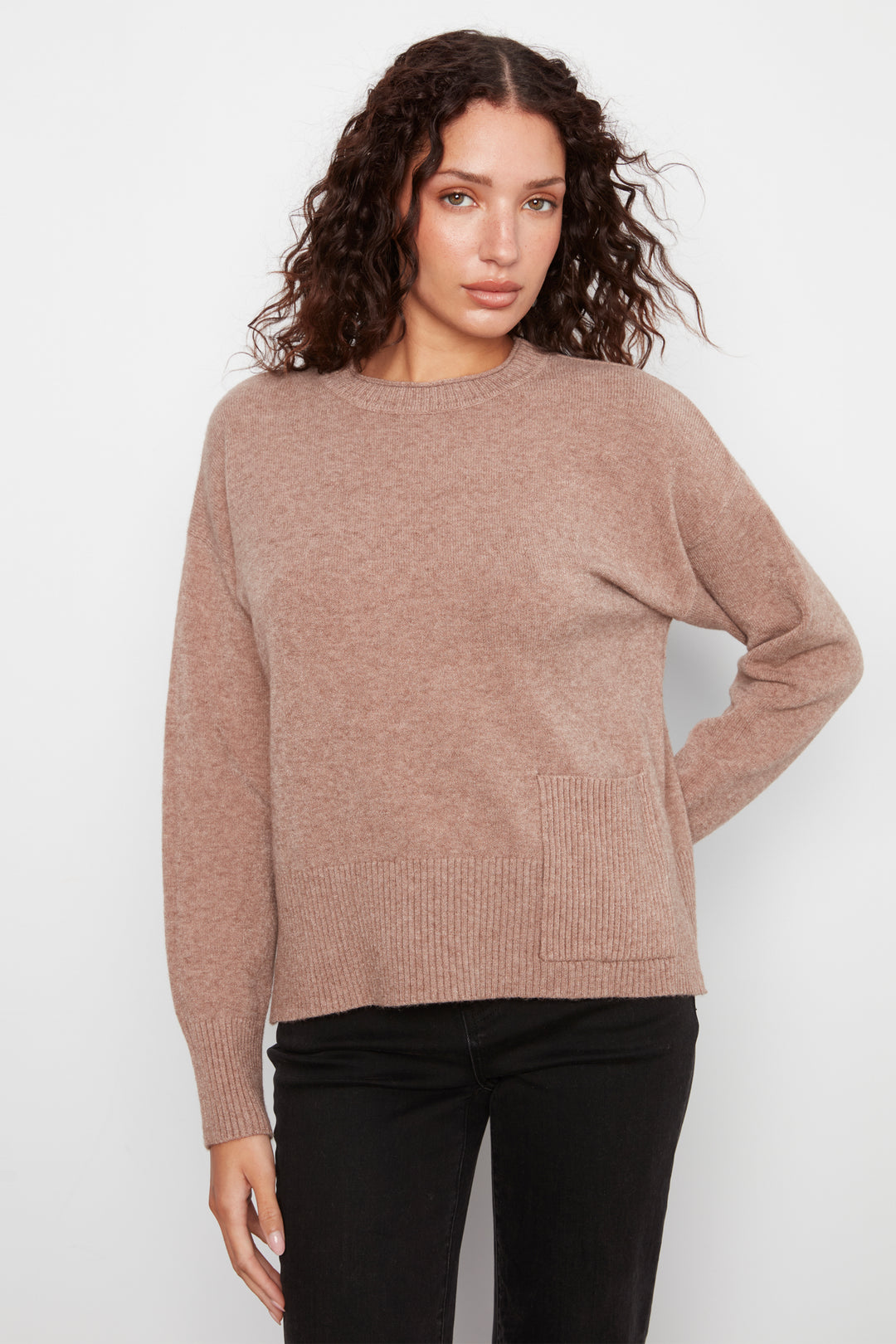 A woman wearing a dusty rose knit crewneck sweater and black pants, standing against a light gray studio background.