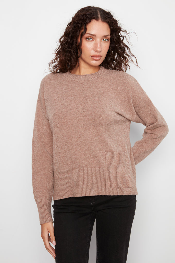 A woman wearing a dusty rose knit crewneck sweater and black pants, standing against a light gray studio background.
