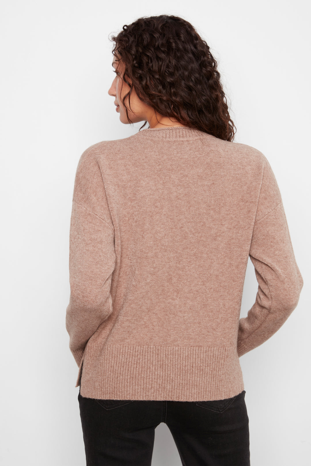A woman with curly brown hair is shown from the back wearing a light brownish-pink knit sweater and black pants against a plain white background.