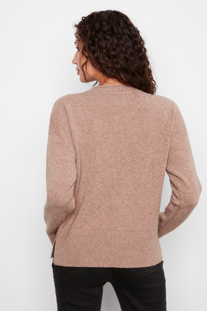 A woman with curly brown hair is shown from the back wearing a light brownish-pink knit sweater and black pants against a plain white background.