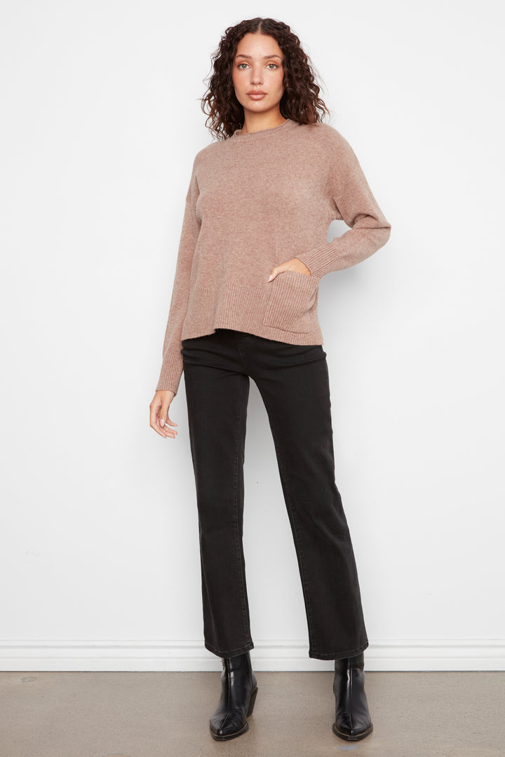 A woman modeling a light brown knit sweater with black trousers and black ankle boots, standing against a white wall.