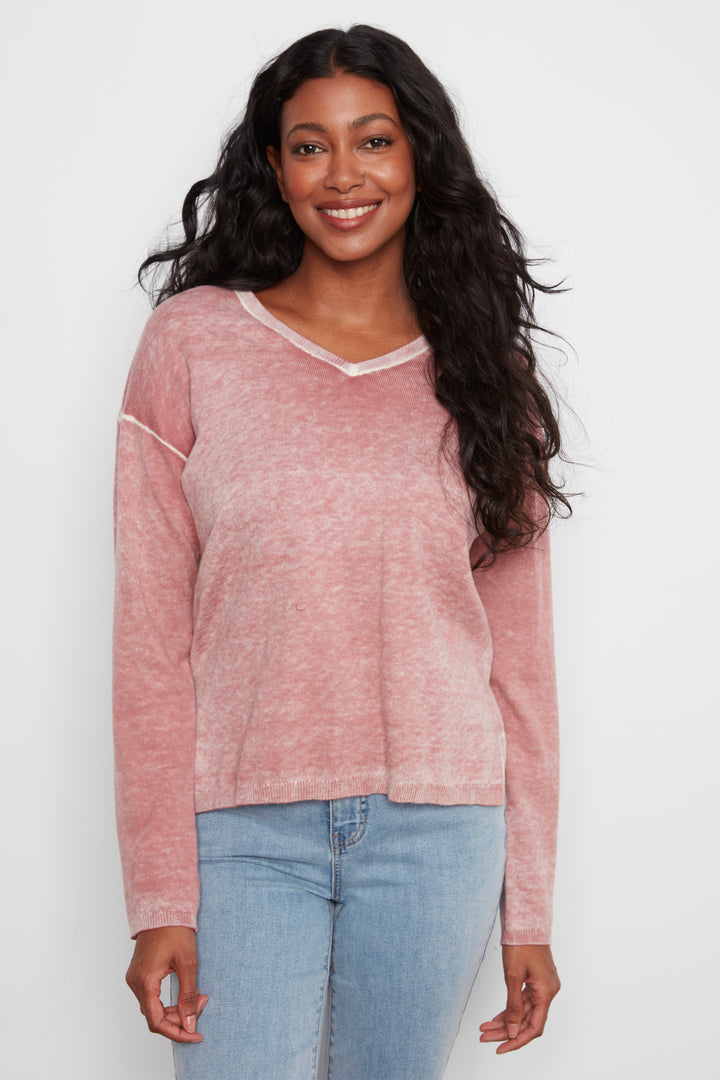 A woman with long, wavy dark hair is wearing a dusty rose pink long-sleeve knit sweater with white contrast stitching and light blue jeans, standing against a plain white studio background.