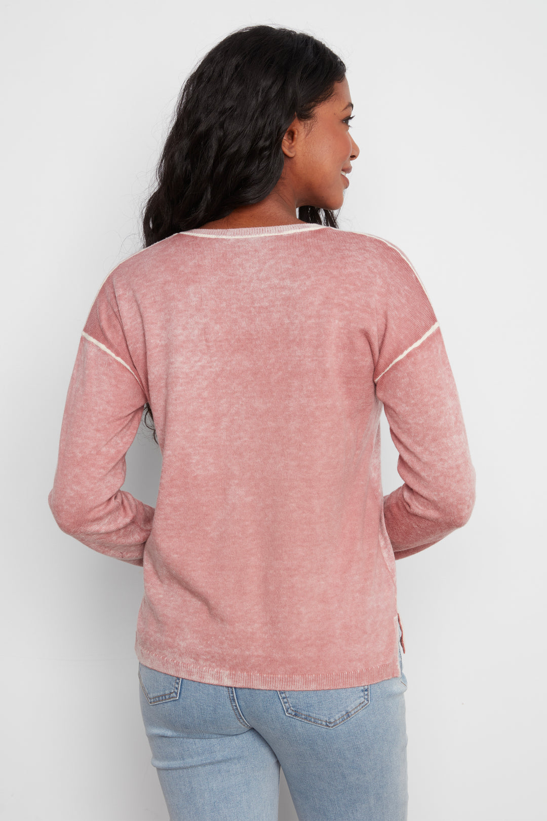 A woman shown from the back wearing a dusty rose long-sleeve top with white contrast stitching details on the shoulders and sides, paired with light blue jeans, against a plain light gray background.