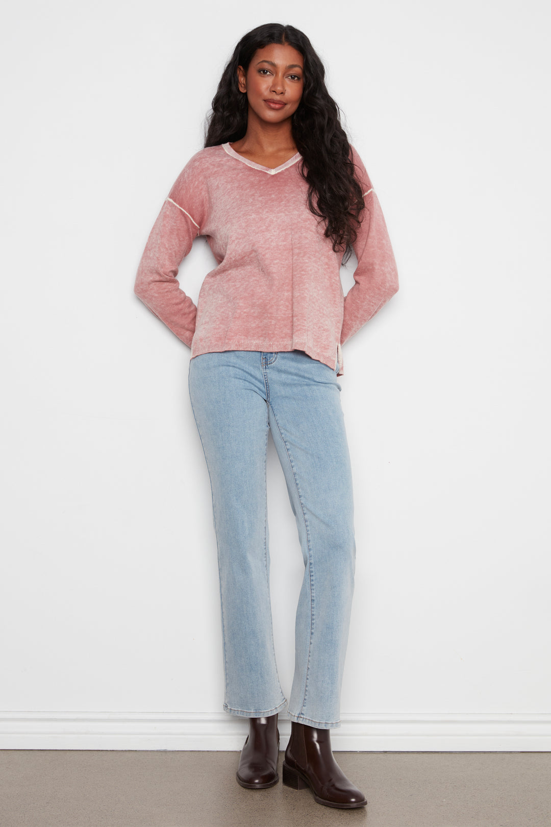 A woman modeling a dusty pink long-sleeve knit sweater with white contrast stitching and a white V-neck trim, paired with light-wash blue jeans and dark brown ankle boots, standing against a white wall.