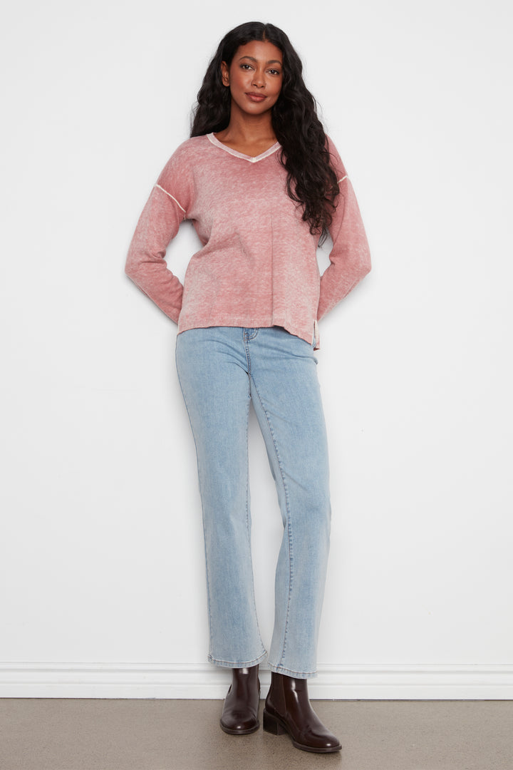 A woman modeling a dusty pink long-sleeve knit sweater with white contrast stitching and a white V-neck trim, paired with light-wash blue jeans and dark brown ankle boots, standing against a white wall.