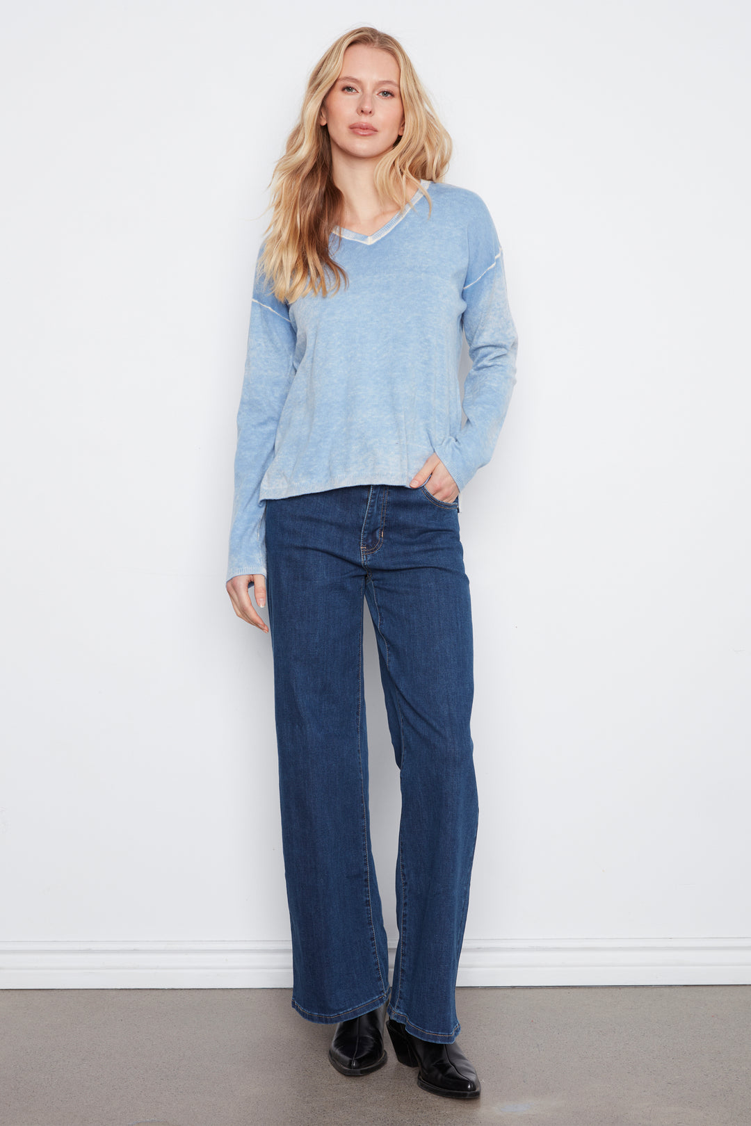 A woman modeling a light blue long-sleeve knit top and dark blue wide-leg jeans against a plain white wall.