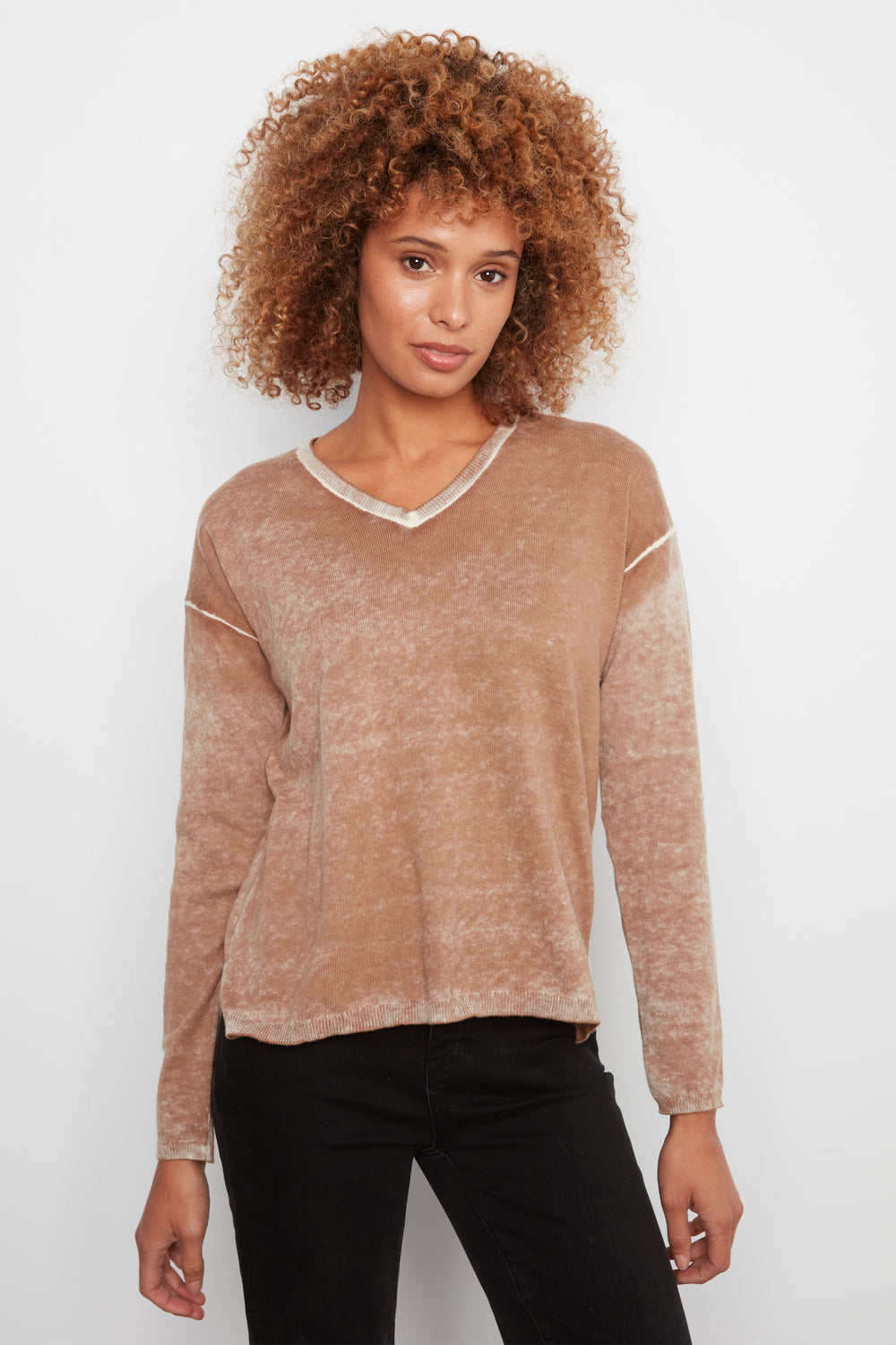 A woman with curly light brown hair wearing a long-sleeve, cropped V-neck knit sweater in a heathered tan-brown color, paired with black pants, against a plain white studio background.