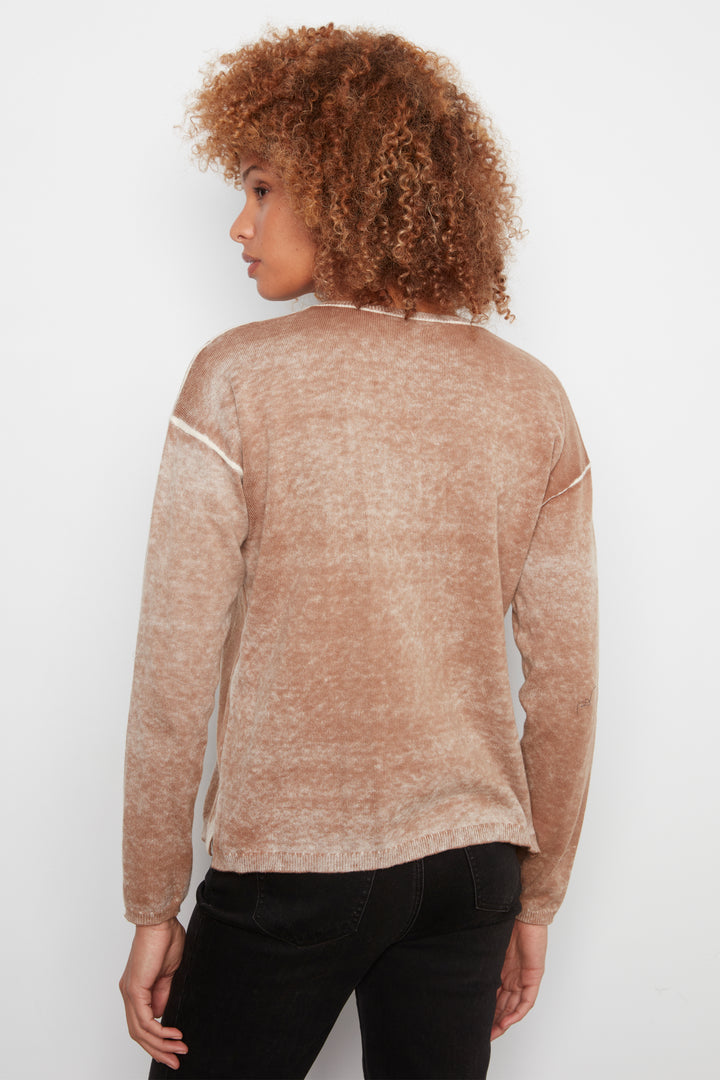 A woman with curly brown hair is shown from the back wearing a marled, washed tan/beige long-sleeve sweater with white contrast stitching on the shoulders, paired with black pants, against a plain white background.