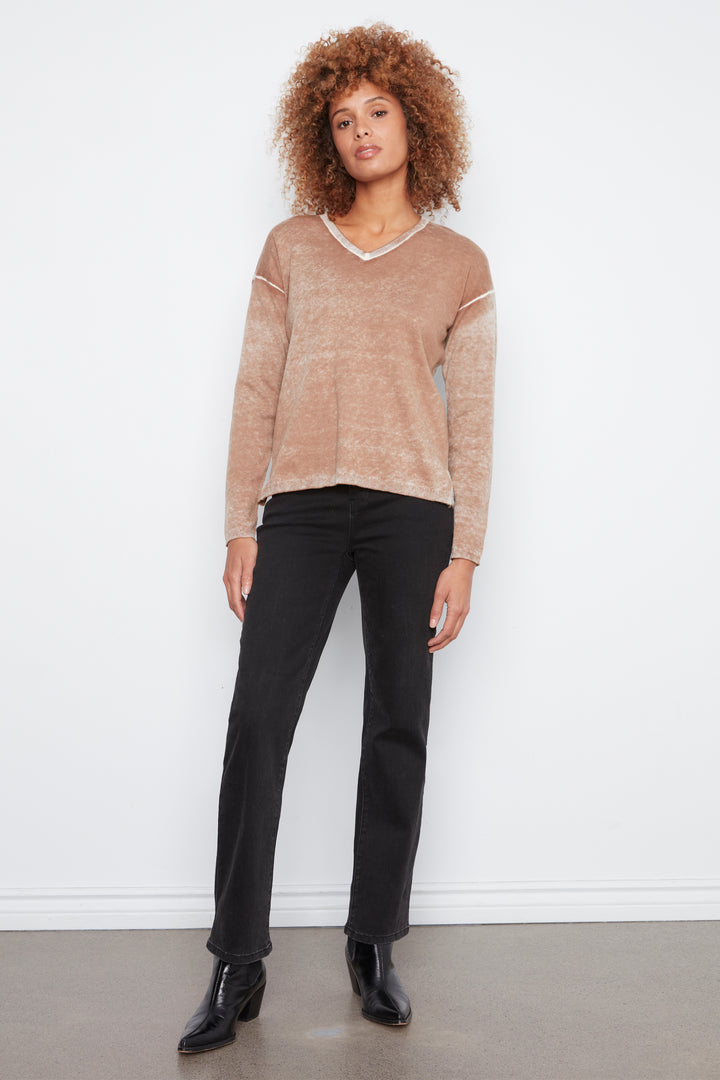 A woman with curly light brown hair stands against a white wall wearing a cropped, marled beige knit sweater with white trim on the V-neck and cuffs, paired with black pants and black ankle boots.