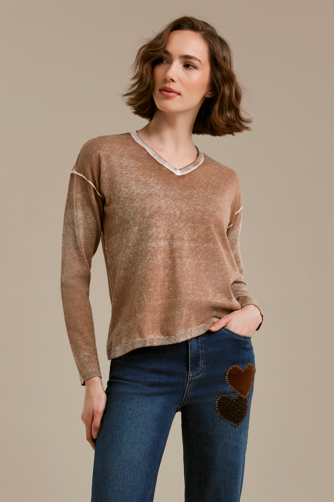 A woman modeling a long-sleeve V-neck knit sweater in a heathered taupe-brown color, featuring white contrast stitching along the shoulders and neckline, paired with blue jeans that have brown heart patches on the pocket.