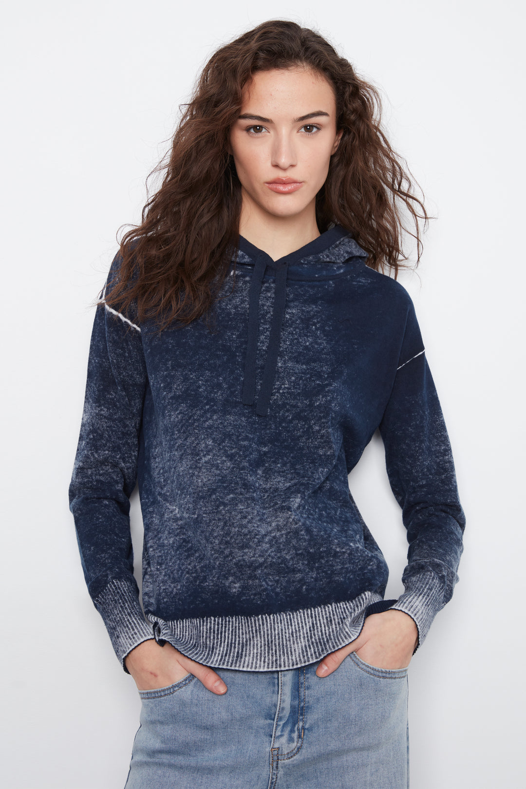 A woman wearing a dark blue acid-wash hooded sweatshirt with ribbed cuffs and hem, paired with light blue jeans, against a plain white studio background.