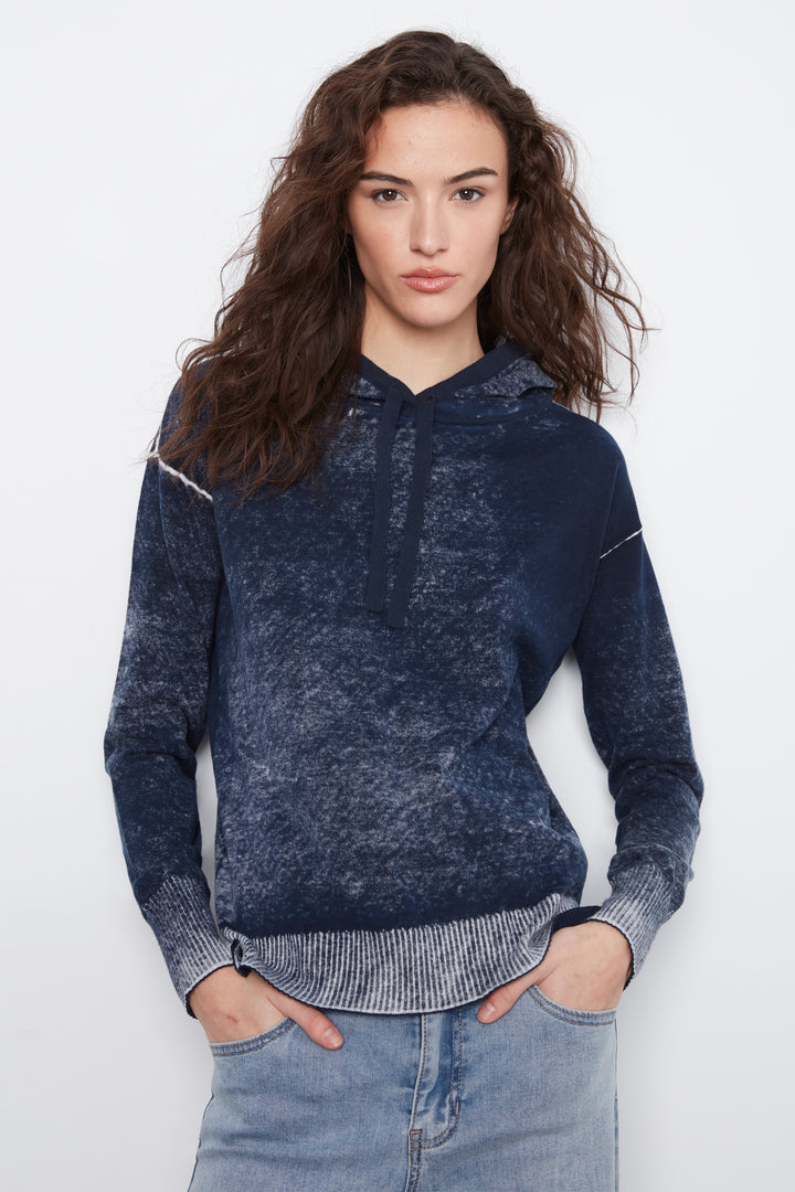 A woman wearing a dark blue acid-wash hooded sweatshirt with ribbed cuffs and hem, paired with light blue jeans, against a plain white studio background.