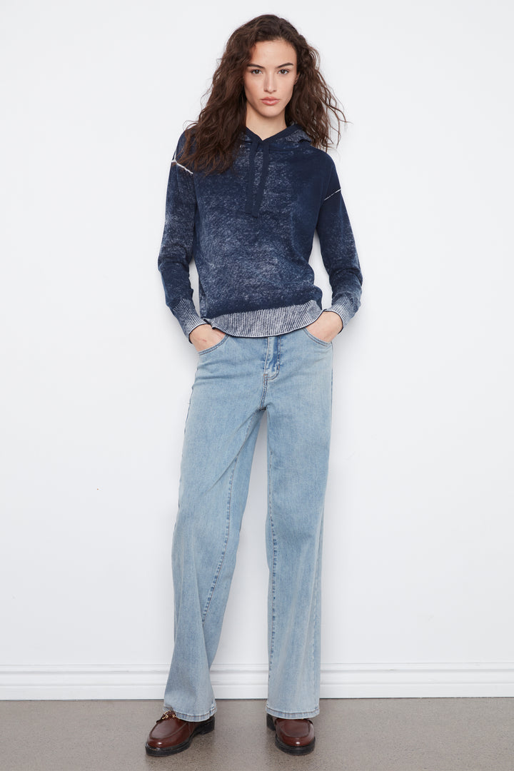 A woman stands against a white wall wearing a dark blue marbled hoodie, light blue wide-leg jeans, and brown leather loafers.