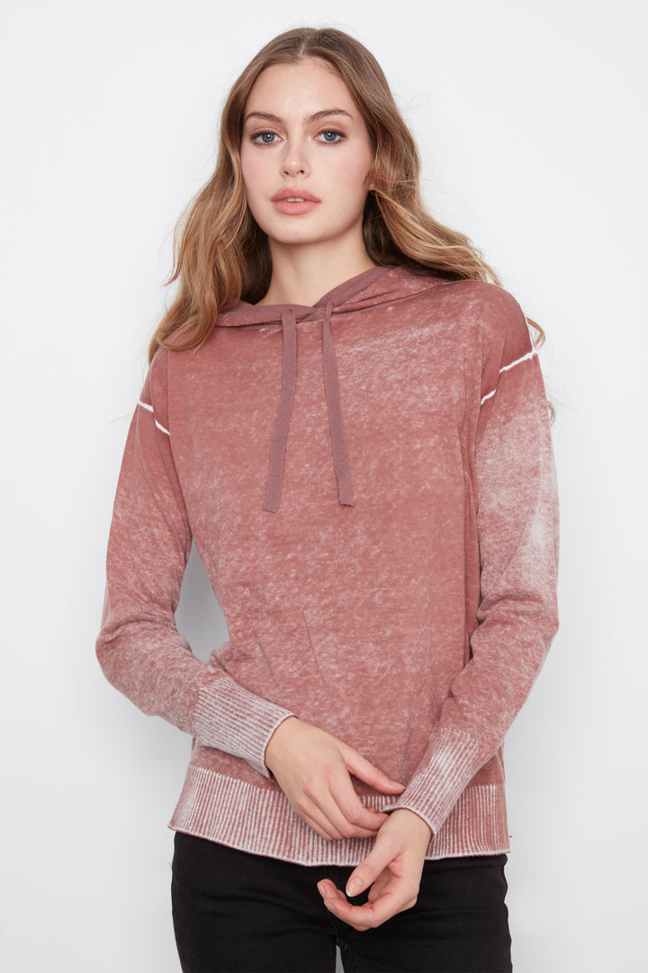 A woman modeling a dusty rose pink hooded knit sweater with a marled texture, featuring drawstrings and white contrast stitching details on the shoulders and cuffs, paired with black pants against a light gray background.