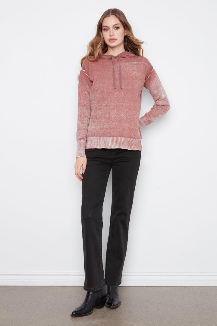 A woman modeling a dusty rose knit hoodie with textured shoulder details and black pants, standing against a plain white wall.