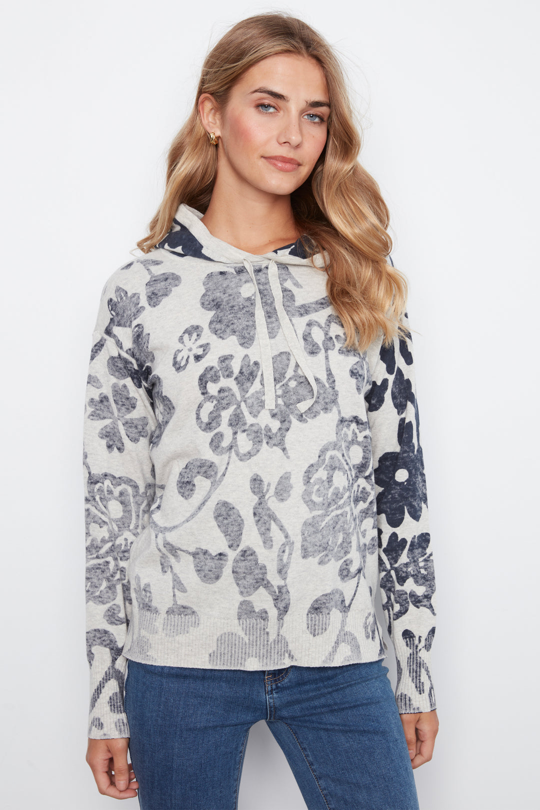 A woman wearing a long-sleeve hooded sweatshirt with a gray and navy floral pattern on a light heather gray background, paired with blue jeans, against a plain white studio background.