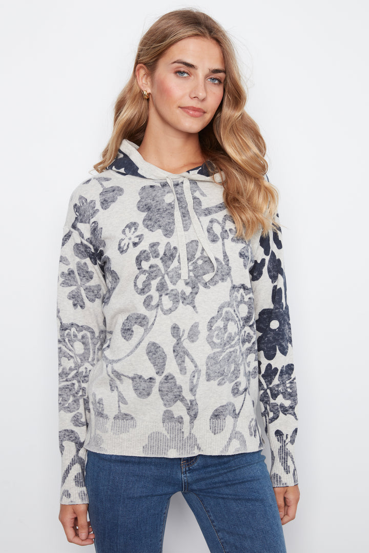 A woman wearing a long-sleeve hooded sweatshirt with a gray and navy floral pattern on a light heather gray background, paired with blue jeans, against a plain white studio background.