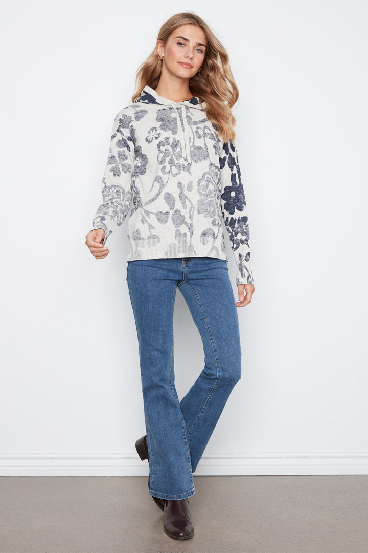 A woman modeling a white long-sleeve hoodie with a gray and navy floral pattern, paired with blue jeans and brown boots, standing against a white wall.