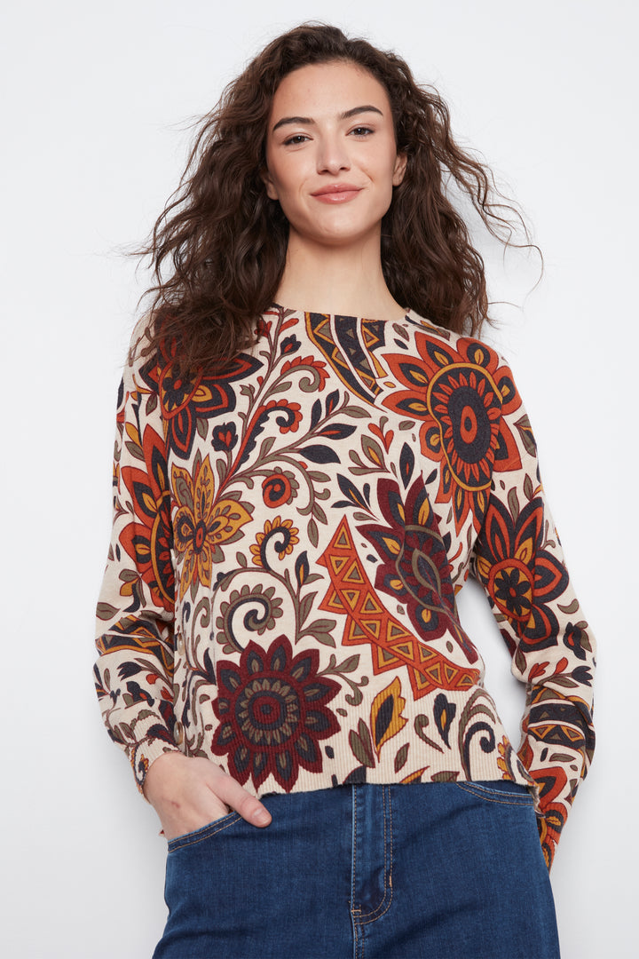 A woman wearing a long-sleeve boat-neck top with a detailed bohemian floral and geometric pattern in rust orange, dark red, brown, and black on a cream background, paired with blue jeans.