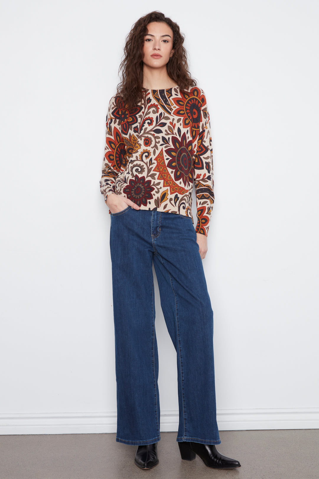 A woman stands against a white wall wearing a long-sleeve V-neck top with a complex floral and geometric pattern in orange, brown, black, and cream, paired with dark blue wide-leg jeans and black ankle boots.