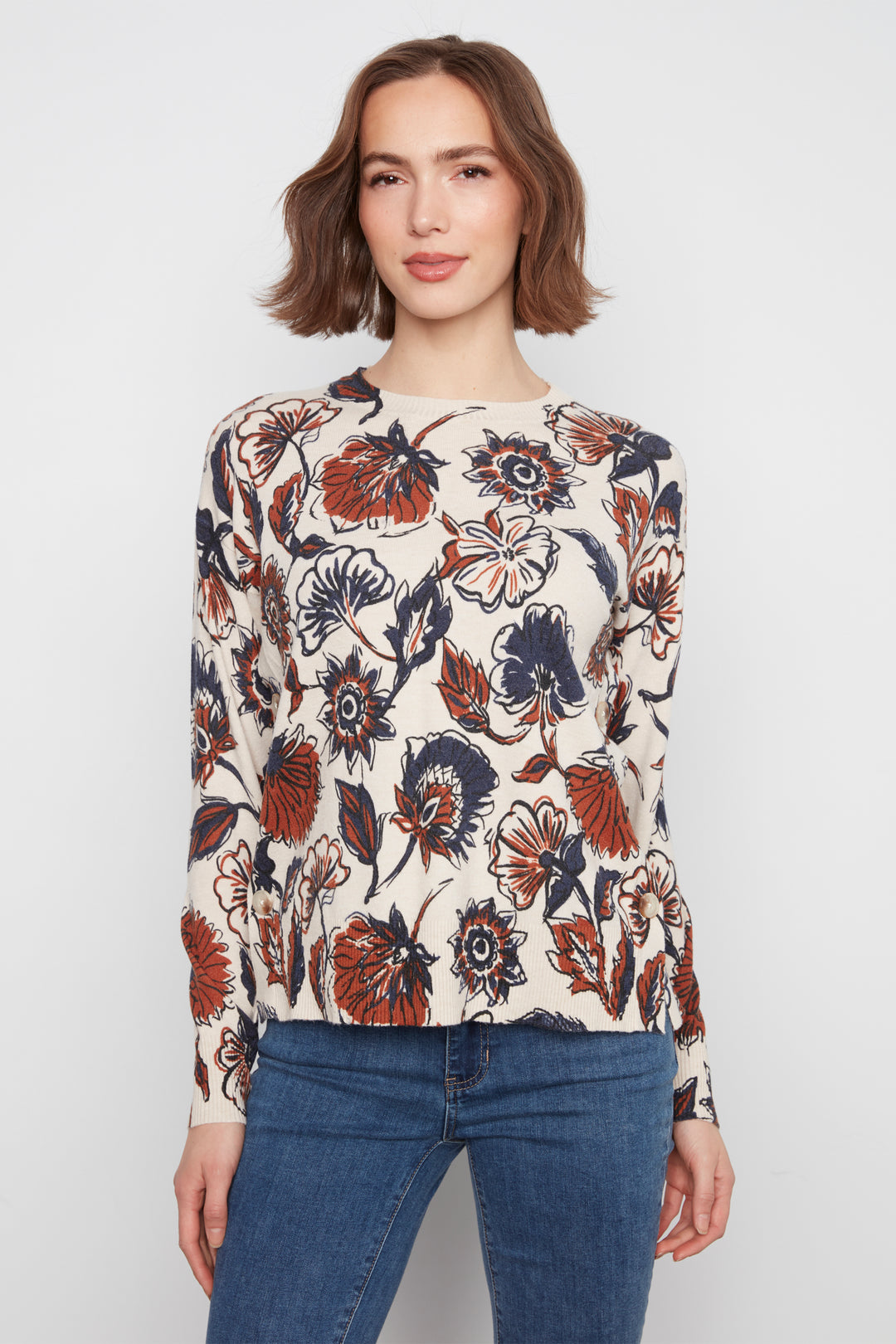 A woman modeling a cream-colored long-sleeve knit top with a bold floral pattern in rust orange, navy blue, and black, paired with blue jeans against a light gray studio background.