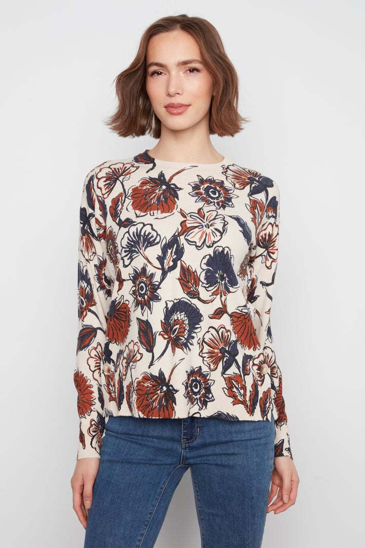 A woman modeling a cream-colored long-sleeve knit top with a bold floral pattern in rust orange, navy blue, and black, paired with blue jeans against a light gray studio background.