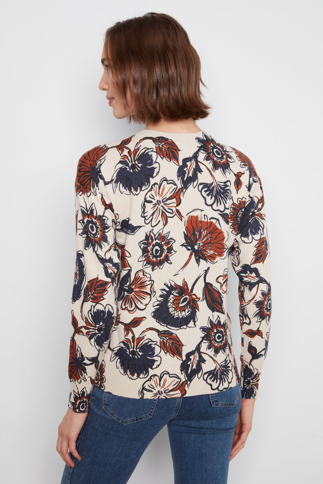 A woman shown from the back wearing a cream-colored long-sleeve top with a bold floral pattern in rust orange, navy blue, and black, paired with blue jeans against a light gray studio background.