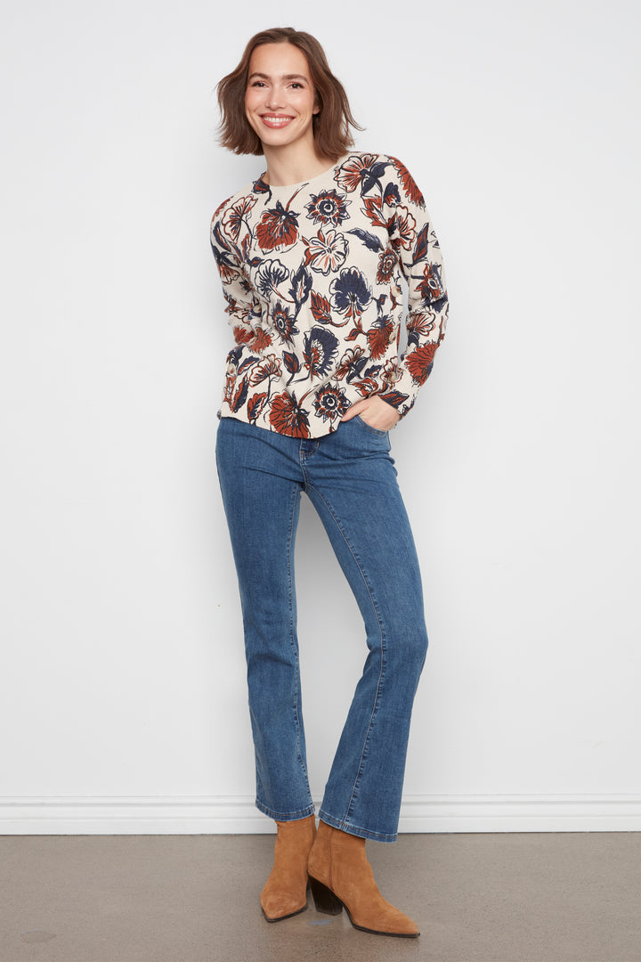 A woman modeling a long-sleeve floral print top with blue jeans and brown suede ankle boots, standing against a plain white wall.