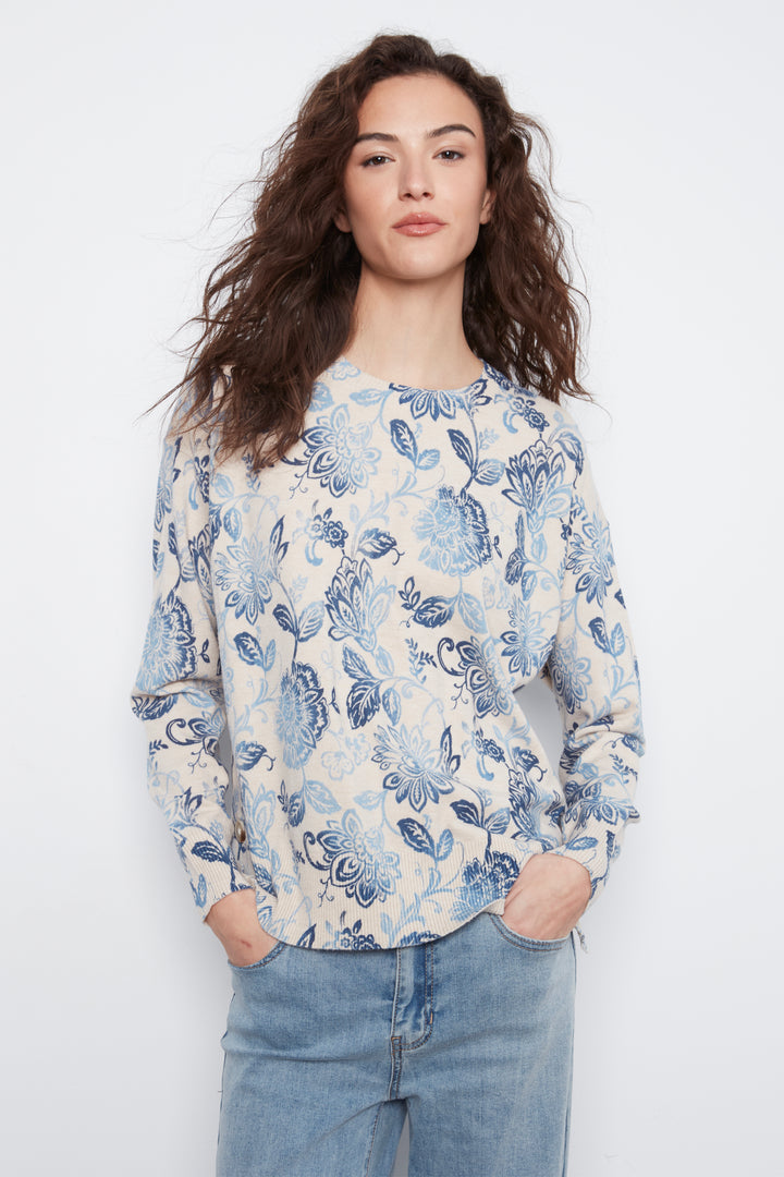 A woman with long, wavy brown hair wearing a cream-colored long-sleeve top with a blue floral pattern, paired with light-wash blue jeans, standing against a plain white studio background.