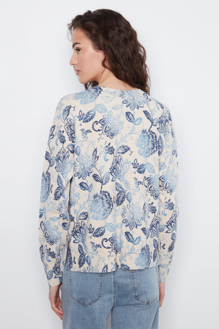 A woman shown from the back wearing a cream-colored long-sleeve blouse with a blue floral pattern, paired with light blue jeans, against a plain white background.