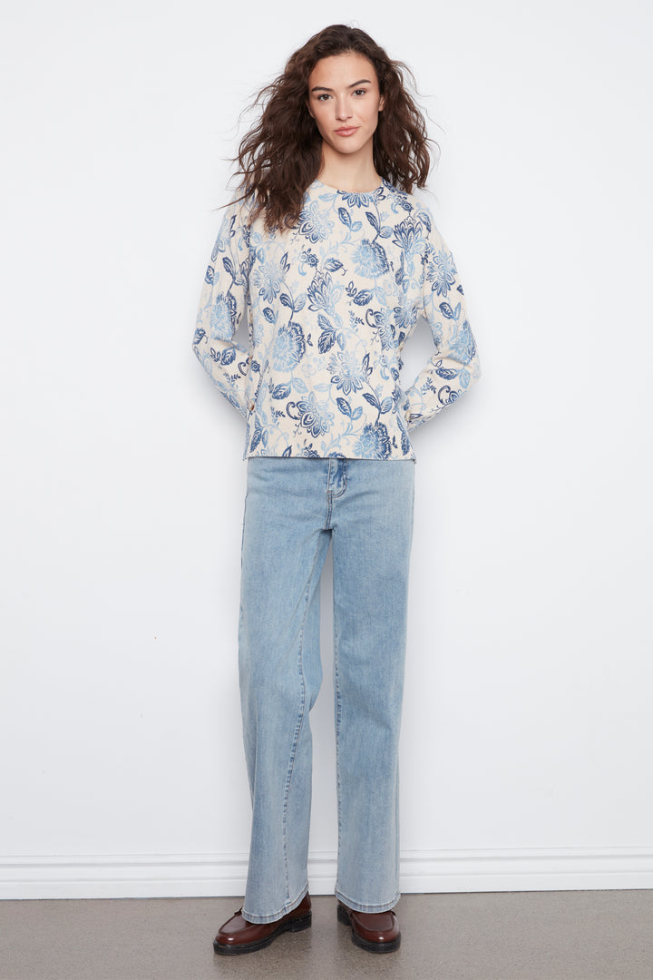 A woman stands against a white wall wearing a cream long-sleeve blouse with a blue floral print, light-wash wide-leg jeans, and dark brown loafers.
