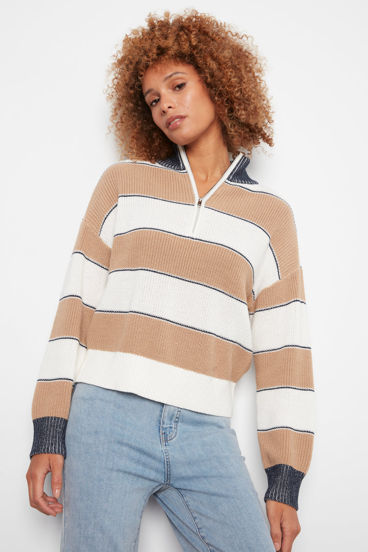 A woman with curly light brown hair wearing a beige, white, and navy striped half-zip sweater and light blue jeans, posing against a plain white studio background.