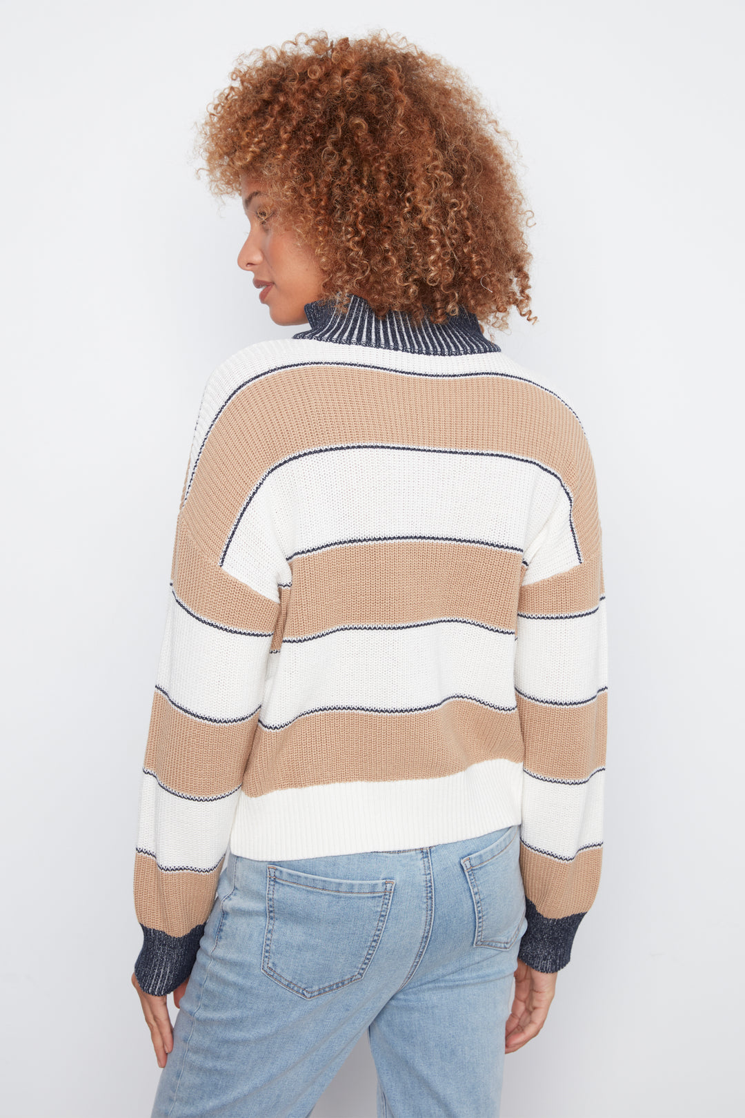 A woman with curly brown hair is shown from the back wearing a beige and white horizontally striped knit sweater with dark navy ribbed trim, paired with light blue jeans, against a plain white studio background.