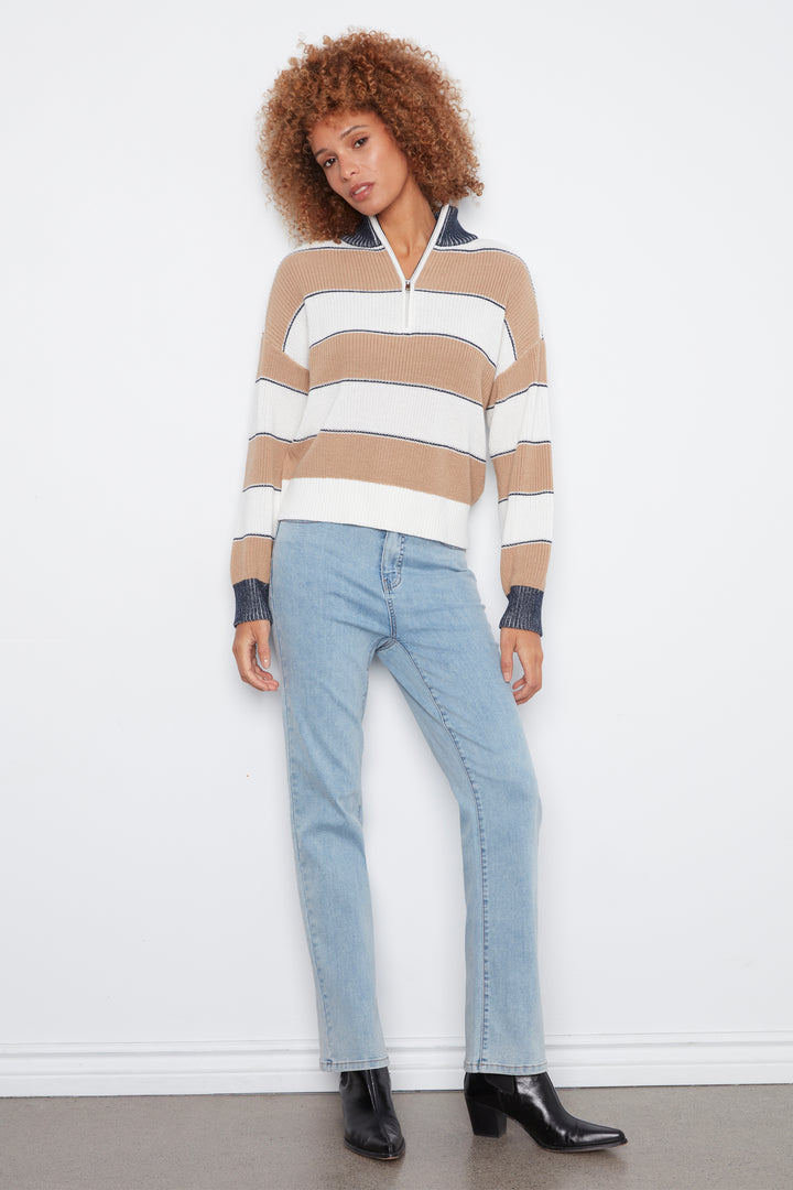 A model with curly brown hair stands against a white wall wearing a beige, white, and navy striped half-zip sweater, light-wash blue jeans, and black ankle boots.