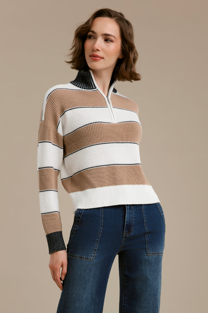 A woman modeling a ribbed knit half-zip sweater with horizontal stripes in beige, white, and dark charcoal, paired with dark blue jeans against a solid beige studio background.