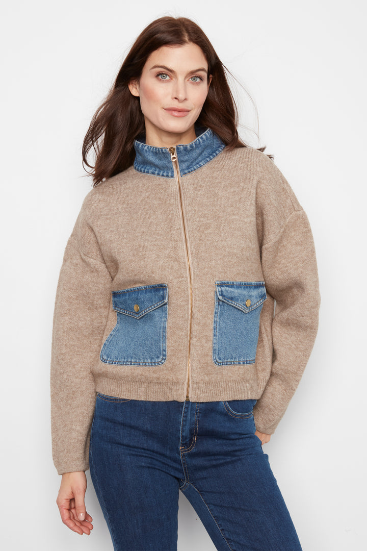 A woman wearing a beige knit zip-up jacket with blue denim collar, pocket, and front patch pockets, paired with blue jeans, against a plain light gray studio background.