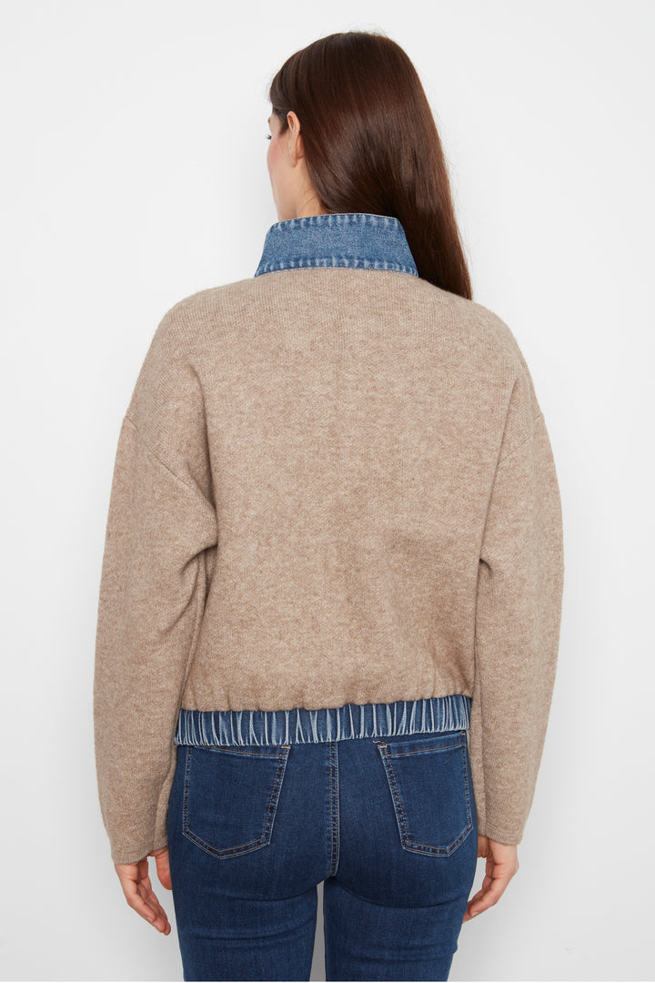 A woman viewed from the back wearing a cropped beige knit sweater with a blue denim collar and hem, paired with dark blue jeans, against a plain white studio background.