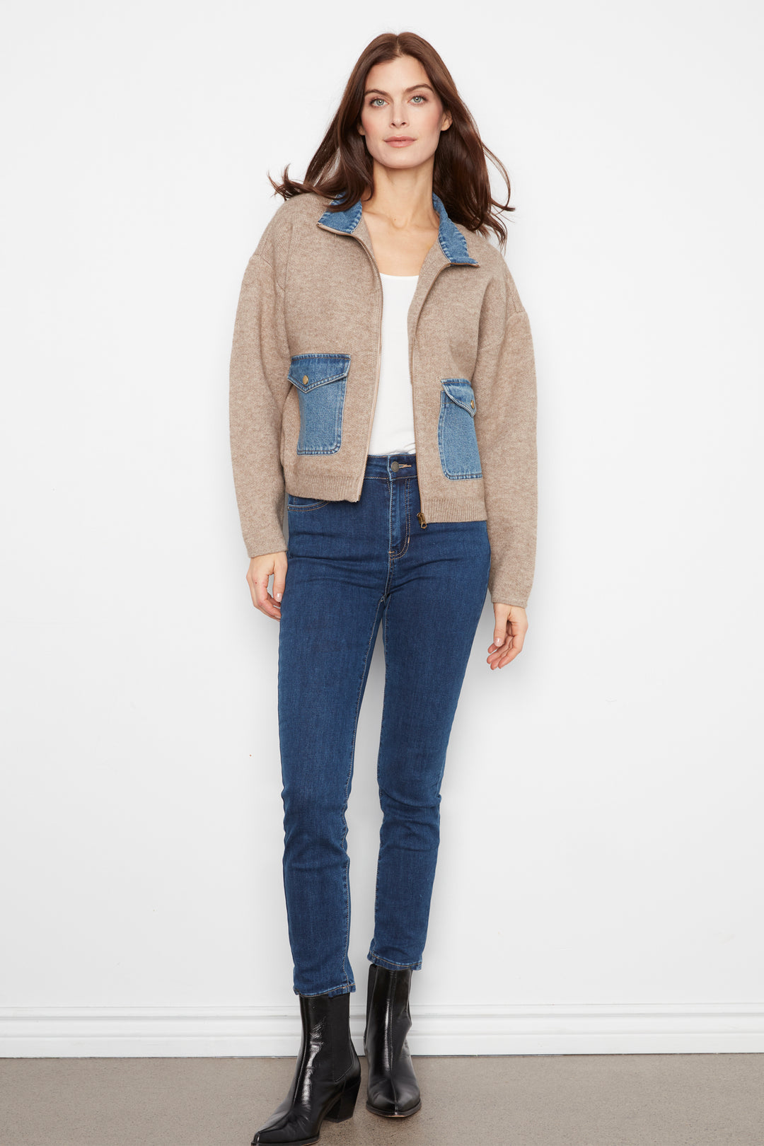 A woman modeling a beige knit zip-up jacket with blue denim pocket and collar details, paired with blue jeans and black ankle boots, standing against a white wall.