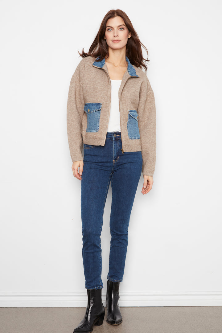 A woman modeling a beige knit zip-up jacket with blue denim pocket and collar details, paired with blue jeans and black ankle boots, standing against a white wall.