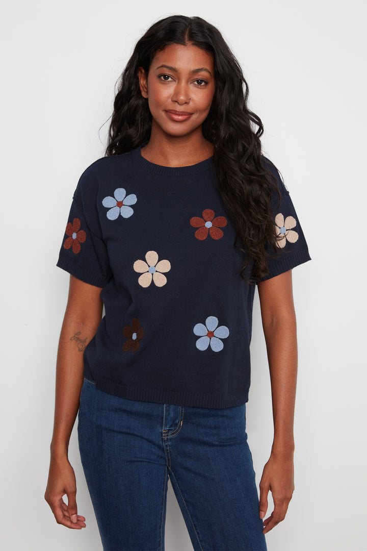 A woman modeling a navy blue short-sleeve knit top with colorful embroidered flowers, paired with blue jeans, against a plain light gray background.